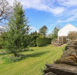 A garden with green grass trees and a stone wall next to a white building at Mell View Cottage in Ullswater