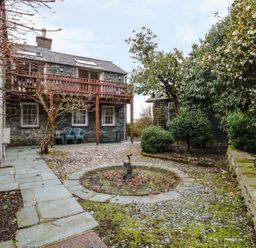 An outdoor area with a stone pathway, trees, bushes, a small fountain, and a stone building with a wooden balcony and plastic chairs at Coombe Cottage in Borrowdale