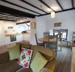 A kitchen with white cabinets and wooden countertops next to a dining area with a wooden round table and four chairs and a brown sofa with green and patterned cushions at Laurel Cottage in Spark Bridge