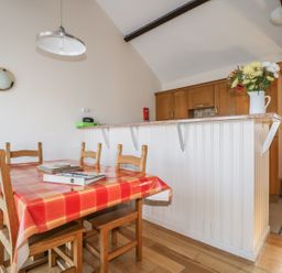 A dining area with a wooden table covered with a red checkered tablecloth and six wooden chairs next to a kitchen counter with a flower vase on top at Esthers Barn in Cockermouth
