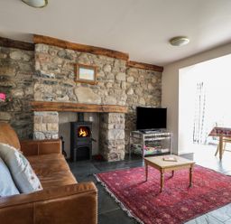 A living room with a leather sofa a stone fireplace with a wood stove a red rug a coffee table a television and a dining table at Beckside in Cockermouth