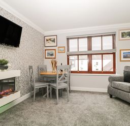 A dining area with a round table and four chairs next to a window and an armchair near a wall-mounted television above a fireplace at Andra Ghyll in Keswick