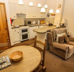 A kitchen with white cabinets a stove microwave and kettle next to a seating area with a brown armchair and a wooden dining table with chairs at Juniper Cottage in Ambleside