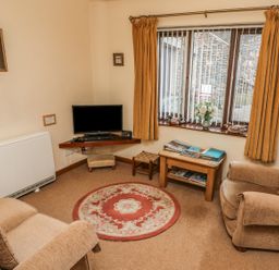 A living room with two armchairs a round rug a small wooden table with books a wall-mounted television and a window with curtains at Juniper Cottage in Ambleside