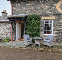 Exterior stone wall of a building with an open white door two gray cushioned chairs and a small round table at The Hayloft in Thirlmere