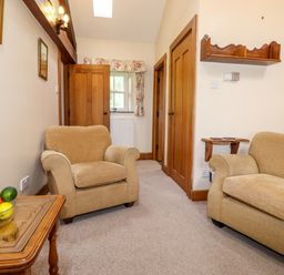A room with two beige armchairs a wooden side table with a bowl of fruit wooden doors and a window with floral curtains at The Hayloft in Thirlmere