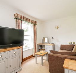 A sitting room with a brown sofa a black TV on a grey cabinet a small wooden table and a window with curtains at Thirlmere Suite in Thirlmere