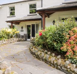 A stone walkway with rock borders leading to a white building entrance with two outdoor chairs and bushes on both sides at Thirlmere Suite in Thirlmere