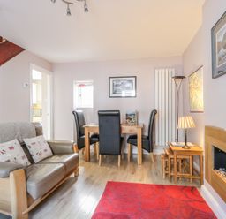A living room with a leather loveseat, black dining chairs at a wooden table, a red rug, wall-mounted fireplace, and staircase at Swinside Cottage in Keswick