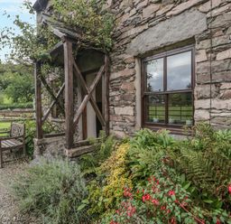 The exterior of a stone cottage with a wooden bench plants and a wooden framed window at Tanner Croft Cottage in Grasmere