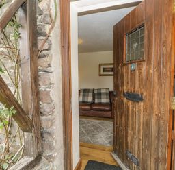 An open wooden front door showing a glimpse of a living room with a brown leather sofa and plaid cushions at Tanner Croft Cottage in Grasmere