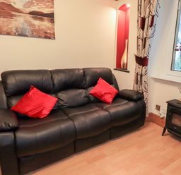 A black leather sofa with two red cushions next to a window and a small black stove at Granary Nook in Windermere