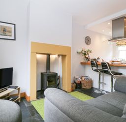 A living room with grey sofas a television a wood stove and a kitchen counter with two bar stools at Banks Place in Keswick