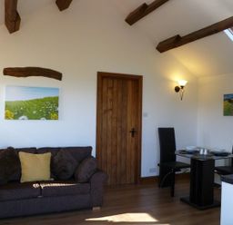 A sitting area with a dark sofa and cushions next to a wooden door and a dining table with two black chairs at Buttercup Cottage in Catterlen