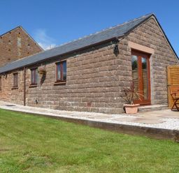 An exterior view of a stone cottage with brown framed windows a grass lawn and a small patio with wooden table and chairs at Buttercup Cottage in Catterlen