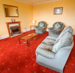 A living room with green upholstered sofa and chairs a glass coffee table a fireplace with a mirror above and a red patterned carpet at Low Skelgill in Troutbeck