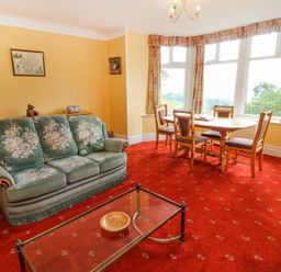 A living room with a green floral sofa a glass coffee table a wooden dining table with four chairs and large windows at Low Skelgill in Troutbeck