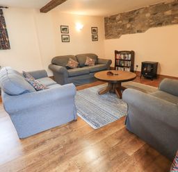 A living room with three sofas a round wooden table a bookshelf and a small stove at Tithe Cottage in Cockermouth