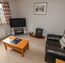 A living room with a brown leather sofa a wooden coffee table a flat screen TV on a stand a basket with books a small stove and a framed picture on the wall at Long Crag Annexe Ambleside
