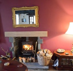 A room with a lit fireplace a basket of firewood a table lamp and a gold framed mirror at Greety Gate House in Foxfield