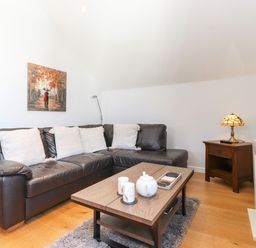 A living room with a black leather sectional sofa with white cushions a wooden coffee table with a teapot and candles a side table with a lamp and a wall-mounted tv at Sky View in Keswick