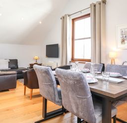 A dining area with a wooden table set for six and grey chairs next to a living room with dark sofas and a wall-mounted TV at Sky View in Keswick