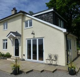 A yellow two-story house with black door and white framed windows a stone patio with two black chairs and potted plants at Eribel Cottage in Threlkeld