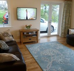 A living room with two sofas a wooden TV stand a wall-mounted TV and large windows at Eribel Cottage in Threlkeld