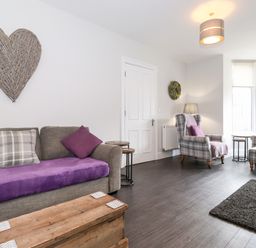 A living room with a grey sofa covered with a purple throw and cushions a wooden chest as a coffee table two plaid armchairs with purple cushions near a window at Latrigg House in Keswick