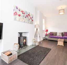 A living room with a wall-mounted television a wood-burning stove a grey sectional sofa with purple cushions and blankets wooden floor and a floral painting above the stove at Latrigg House in Keswick
