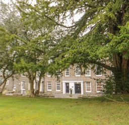 A stone building with multiple windows behind large trees and a grassy lawn at Derwentwater Apartment in Keswick