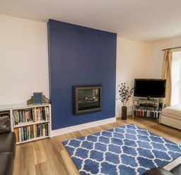 A living room with a blue patterned rug a blue accent wall with a fireplace bookshelves leather sofas a glass TV stand and a window with curtains at Derwentwater Apartment in Keswick