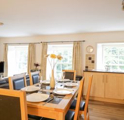 A dining area with a wooden table set with plates and glasses next to a kitchen with light wood cabinets and windows at Derwentwater Apartment Keswick