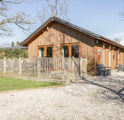 A wooden house with a porch and chairs on a gravel driveway surrounded by trees at Holmedell in Greystoke