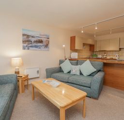 A living room with sofas pillows and a wooden coffee table next to a kitchen with cabinets at Treetops in Windermere