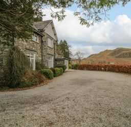 A stone house with bushes in front and a gravel driveway with hills in the background at The Old Vicarage in Keswick