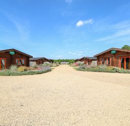 Four wooden lodges with gravel driveway and garden beds under a blue sky at Ash Lodge in Greetham Rutland