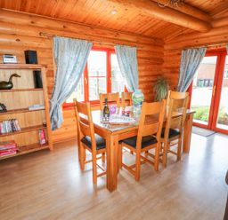 A dining area with wooden table and chairs next to windows with blue curtains and a wooden shelf with books and board games at Yew Lodge in Greetham Rutland