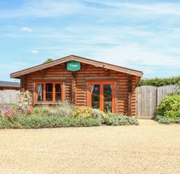 A wooden cabin with orange window frames and a green sign reading Yew surrounded by plants and gravel in front at Yew Lodge in Greetham Rutland