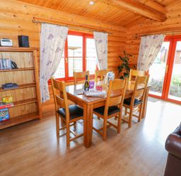A dining area with a wooden table and six chairs near windows and glass doors with floral curtains in a log cabin at Willow Lodge in Greetham Rutland