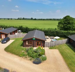 Three wooden lodges with fenced patios and parked car on gravel drive with green fields and trees at Sycamore Lodge in Greetham Rutland