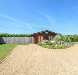 A gravel driveway leading to a wooden cabin with a garden and a fence in the background at Cedar Lodge in Greetham Rutland