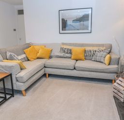 A grey corner sofa with yellow and grey cushions and a framed picture above at Wynding Apartment in Amble-By-The-Sea