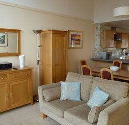 A living area with a beige sofa and light blue cushions a wooden cabinet with a white vase and a stereo a floor lamp a dining table with chairs and a kitchen with wooden cabinets at 16 Keswick Bridge in Keswick
