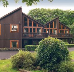 A brown wooden building with large windows and balcony surrounded by green bushes and grass at 20 Keswick Bridge in Keswick