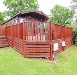 A wooden cabin with a large deck and railing in a grassy area with trees at 10 Thirlmere White Cross Bay near Troutbeck Bridge