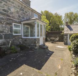 An outdoor stone patio area with a bench a small porch door a garden shed table and chairs and various plants at Ty Uchaf in Dyffryn Ardudwy