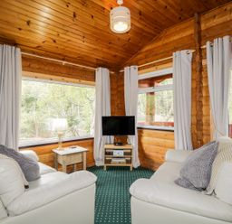 A sitting area with two white armchairs a small wooden table with a lamp a TV on a stand and large windows at Eagle Lodge in Strathpeffer
