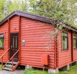 A red wooden cabin with a dark door and windows surrounded by trees at Otter Lodge in Strathpeffer