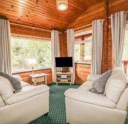 A living room with two cream sofas a TV on a stand a side table with a lamp and large windows at Otter Lodge in Strathpeffer